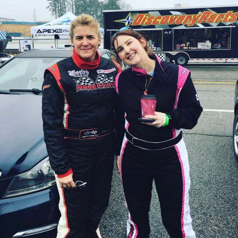 Two women in race suits by car and Discovery Parts trailer.